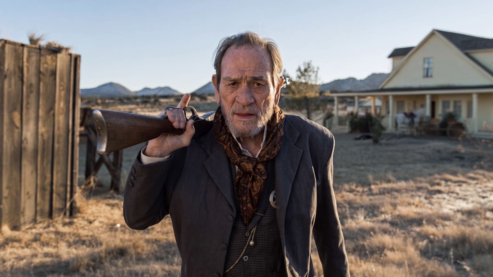 Tommy Lee Jones as the marshal in a gray suit and patterned neckerchief, a rifle over his shoulder, standing in dry grass in front of a ranch house