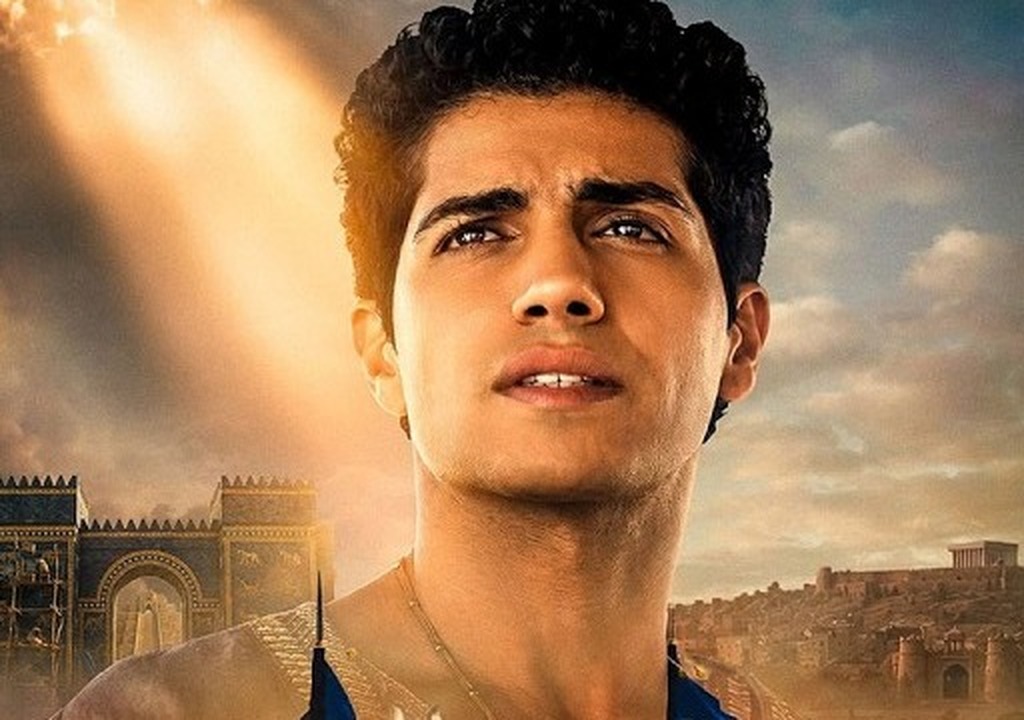 Mena Massoud as Daniel looks upward in a blue and gold robe before a blue Ishtar Gate and a hillside city in Daniel and the Fiery Furnace