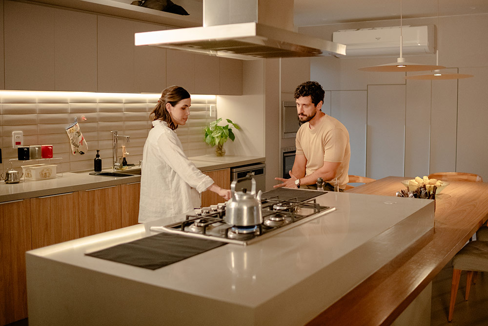 Mario Bregieira, in a tan shirt, sits at a kitchen island facing a woman in a white shirt who stands at a lit gas stove in Flywheel: Ignition of the Soul