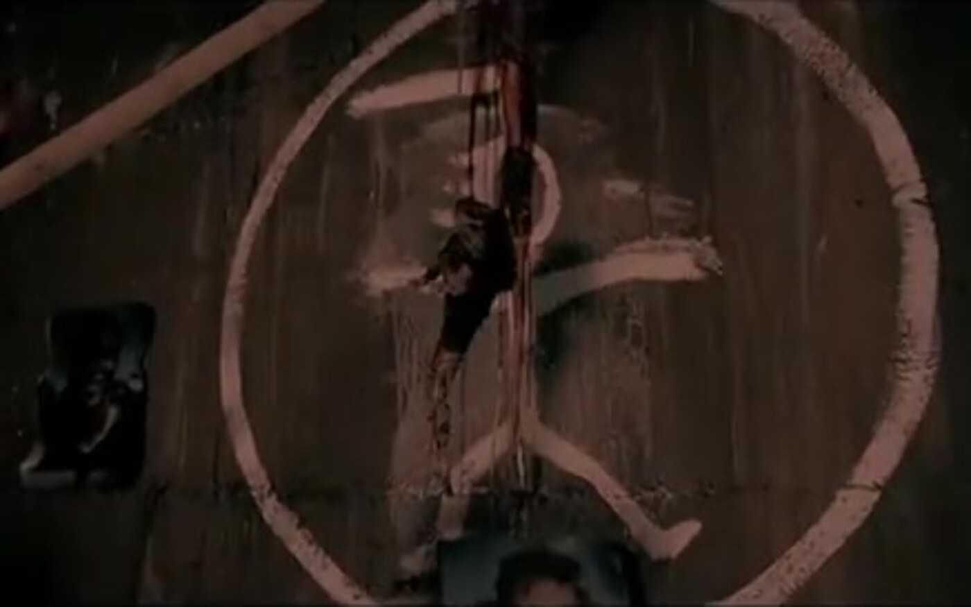 A white ritual symbol painted on a dark wall with red drips and taped photographs in Hangman’s Curse