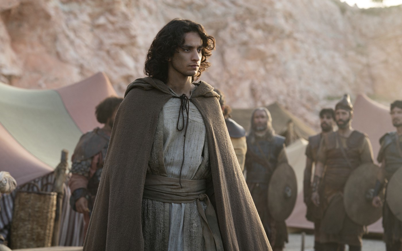 Michael Iskander as David in a brown hooded cloak standing in a desert camp, armored men with shields behind him, in House of David