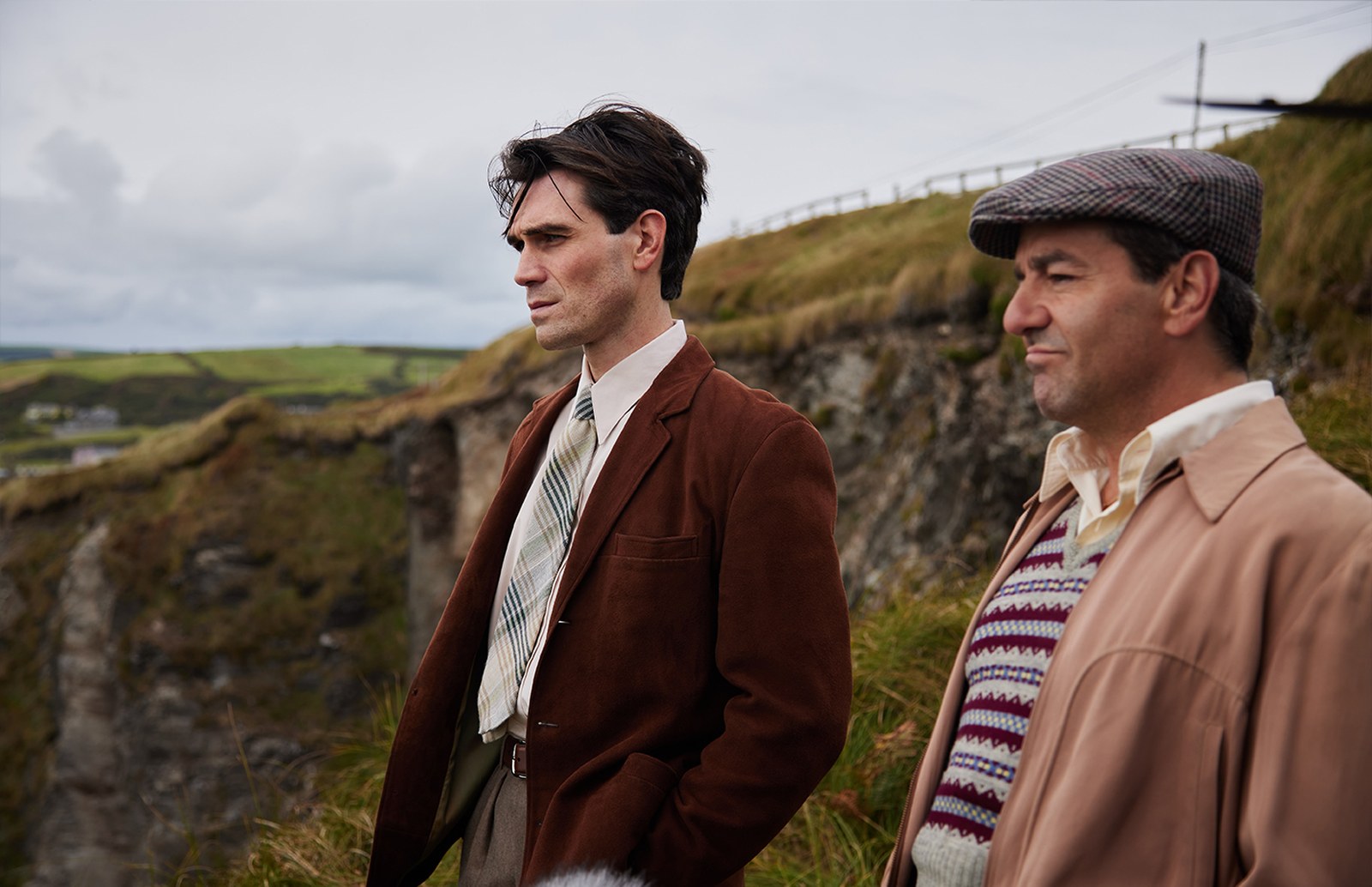 KJ Apa in a brown corduroy jacket and tie stands beside a man in a Fair Isle vest and flat cap on a grassy cliff overlooking green hills