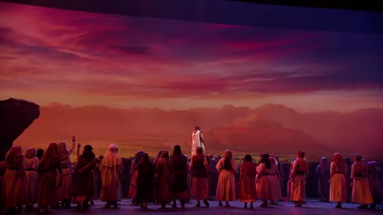 A crowd in robes stands on a stage facing a man in white, with a painted desert and a walled city behind him in Sight & Sound’s Joshua