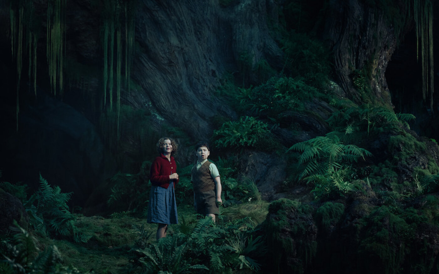 Beatrice Campbell as Polly Plummer in a red cardigan and David McKenna as Digory Kirke in a brown vest stand among ferns in a dark forest in Narnia: The Magician’s Nephew