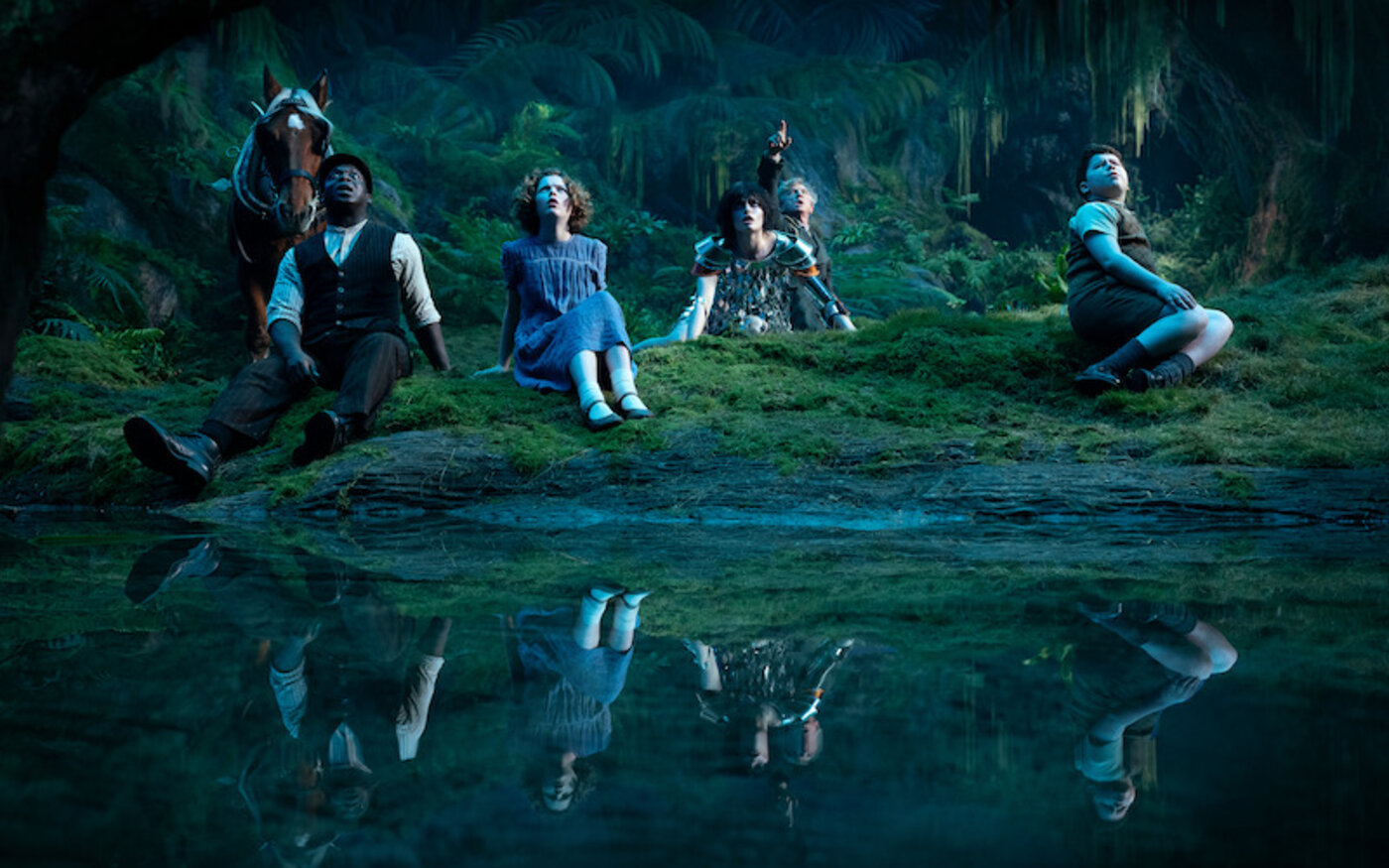 Digory, Polly, and companions sit on a mossy bank beside dark water in a glowing forest in Narnia: The Magician’s Nephew