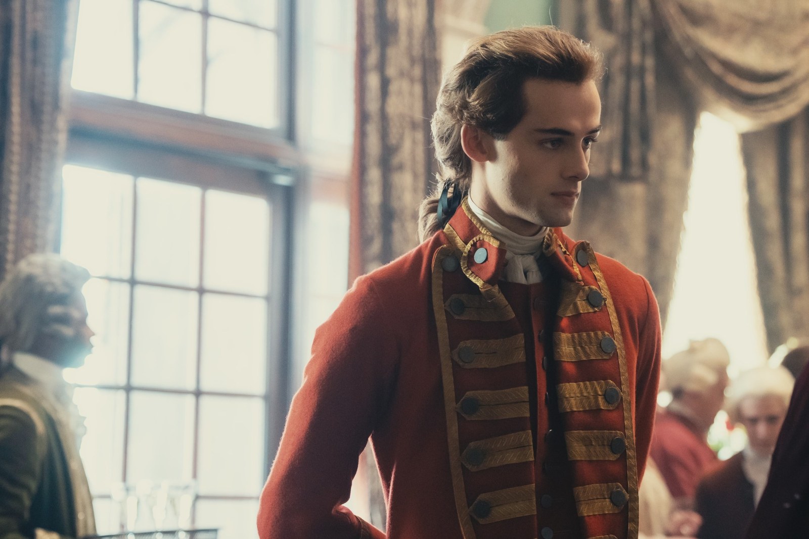 William Franklyn-Miller as young George Washington in a red coat with gold braid and white cravat, standing in a sunlit period interior